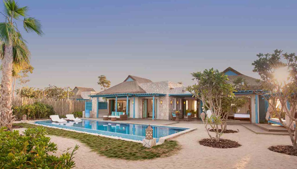 A luxurious, thatched-roof villa featuring a private pool and sandy surroundings is depicted at sunset, highlighting a perfect setting for romantic things to do in Qatar.